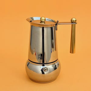 May include: Shiny stainless steel stovetop espresso maker with a gold-colored handle and lid accents. The coffee pot has a bulbous base and a cylindrical upper chamber. The background is a solid orange color.