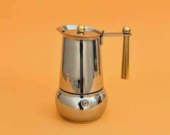 Könnte beinhalten: Glänzender Espressokocher aus Edelstahl mit goldfarbenem Griff und Deckelakzenten. Der Kaffeezubereiter hat einen bauchigen Boden und eine zylindrische obere Kammer. Der Hintergrund ist einfarbig orange.