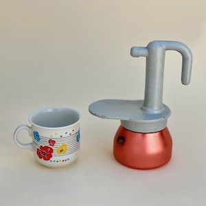 Puede incluir: Una cafetera italiana de color cobre con un asa gris y una taza de cerámica blanca. La taza tiene un estampado floral con flores rojas, azules y amarillas y un asa blanca. La cafetera está al lado de la taza.