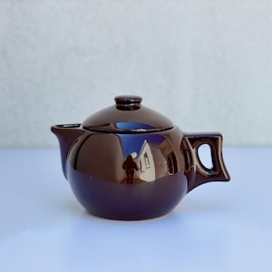 Può includere: Piccola teiera in ceramica marrone scuro con coperchio abbinato. Ha un corpo arrotondato, un beccuccio e un manico con apertura rettangolare. La superficie lucida riflette l'ambiente. Design classico per servire il tè.