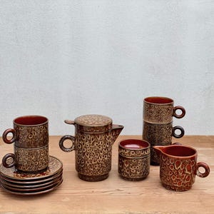 Può includere: Un set da caffè in ceramica marrone con un motivo strutturato. Il set comprende tazze, piattini, una lattiera, una zuccheriera e una caffettiera. I pezzi sono disposti su una superficie di legno.