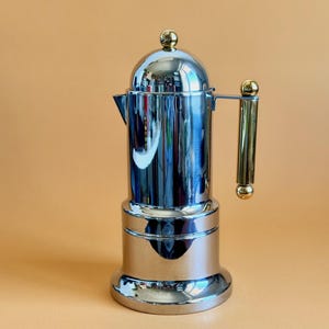 May include: A polished chrome stovetop espresso maker with a rounded top and a gold-coloured handle. The coffee pot has a classic design and is made of polished metal. The handle and top knob are gold-coloured.