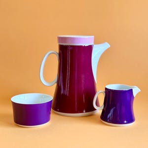 Pode incluir: Um conjunto de chá de cerâmica de três peças em tons de roxo e branco. O conjunto inclui um jarro alto e cilíndrico com uma alça e bico brancos, um pequeno creme e uma tigela redonda. O jarro e o creme são roxos escuros.