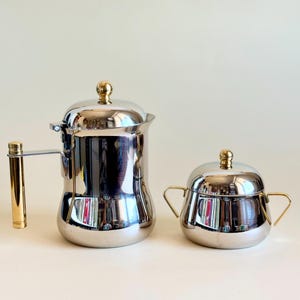 May include: A polished silver-tone metal coffee pot with a gold-tone cylindrical handle and a matching sugar bowl with two gold-tone handles. Both pieces feature a rounded body, a domed lid, and a small gold-tone spherical knob on top.