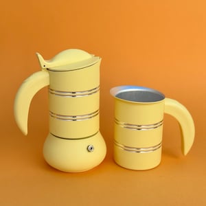 Pode incluir: Uma cafeteira italiana amarelo-pálido com uma jarra de leite a condizer. Ambos os itens apresentam um corpo cilíndrico com duas faixas metálicas horizontais prateadas, uma pega curva e um acabamento mate liso nas superfícies exteriores.