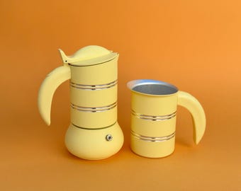 May include: A pale yellow stovetop espresso maker with a matching milk frother pitcher. Both items feature a cylindrical body with two horizontal silver-toned metallic bands, a curved handle, and a smooth matte finish on the exterior surfaces.