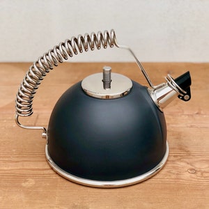 May include: A matte black stovetop kettle with a silver rim and a unique coiled handle. The spout is black, and the lid and handle accents are silver. The kettle sits on a wooden surface.