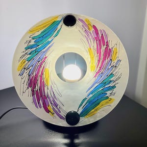 May include: A round, off-white table lamp with a colorful feather design in yellow, blue, pink, and purple. The lamp has a black base and a central light source. The lamp is on a black surface.