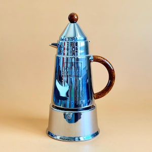 Puede incluir: Una cafetera italiana brillante de acero inoxidable, con un asa marrón y un botón a juego en la parte superior. La cafetera tiene un diseño clásico y está sobre un fondo neutro.