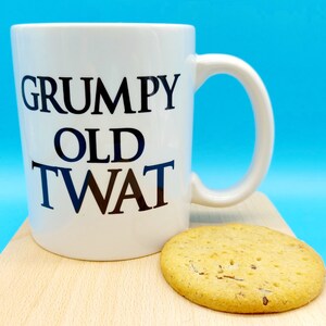 May include: White ceramic mug with black text that reads "Grumpy Old Twat". A biscuit is sitting on a wooden surface in front of the mug.