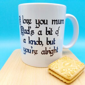 May include: White ceramic mug with a black and blue inscription that reads "I love you mum Dad's a bit of a knob, but you're alright". The mug is sitting on a wooden surface next to a stack of two biscuit-like cookies.