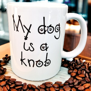 May include: White ceramic coffee mug with black text that reads "My dog is a knob" with paw prints. The mug is sitting on a surface covered in coffee beans.
