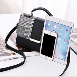 May include: A black and white plaid handbag with a black leather flap and gold hardware. The bag has a top handle and a long shoulder strap. A tablet and a smartphone are placed next to the bag.