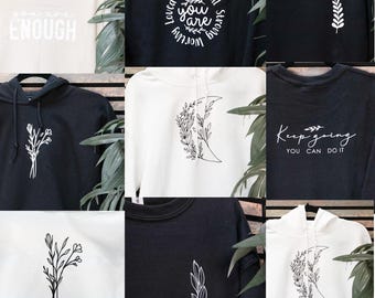 May include: Collection of sweatshirts and hoodies in various colors, including cream, black, and white. Each garment features unique minimalist designs and inspirational text such as "You are Enough", "Beautiful, You are, Strong", and "Keep Going, You Can Do It".