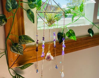 May include: A geometric gold-coloured metal air plant holder with a plant inside, suspended from a chain. Decorative chains with purple and clear beads and crystals dangle below. The background includes a window and green plants.