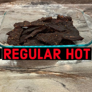 May include: A glass baking dish filled with dark brown beef jerky. The dish is on a wood surface. The text "REGULAR HOT" is printed in red letters on a black background.