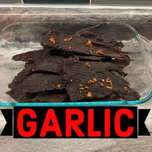 May include: A glass dish filled with dark brown beef jerky with a light brown garlic seasoning. The dish is on a wooden surface with the word "GARLIC" in red letters on a black background.