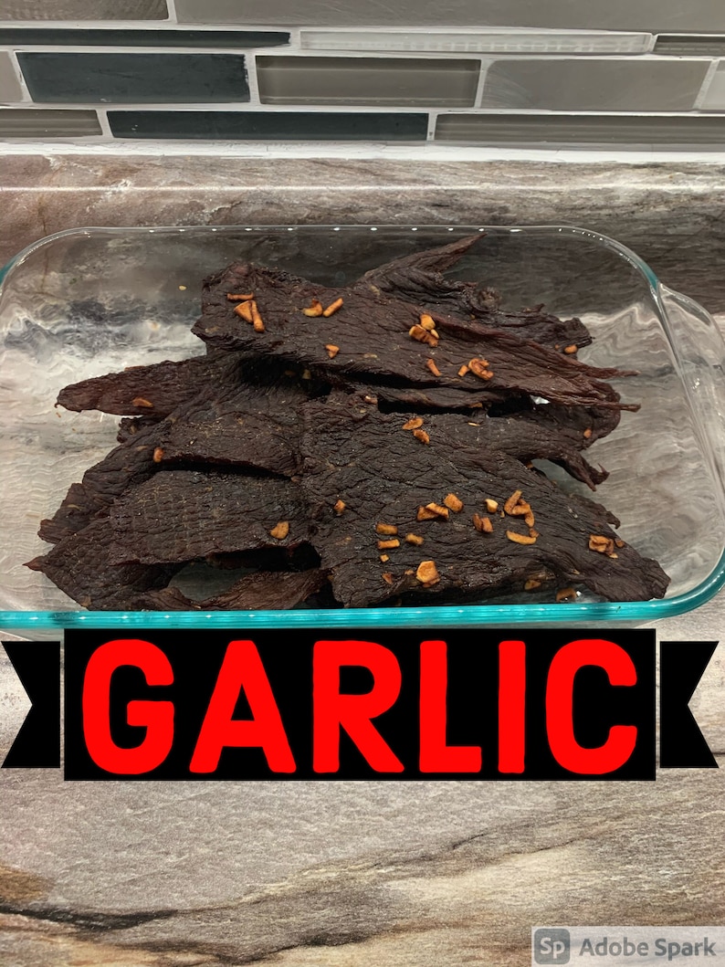 May include: A glass dish filled with dark brown beef jerky with a light brown garlic seasoning. The dish is on a wooden surface with the word "GARLIC" in red letters on a black background.