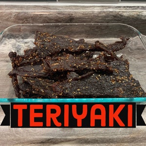 May include: A glass dish filled with dark brown, chewy, teriyaki beef jerky with sesame seeds. The dish is on a wooden surface with the word "TERIYAKI" in red letters on a black background.