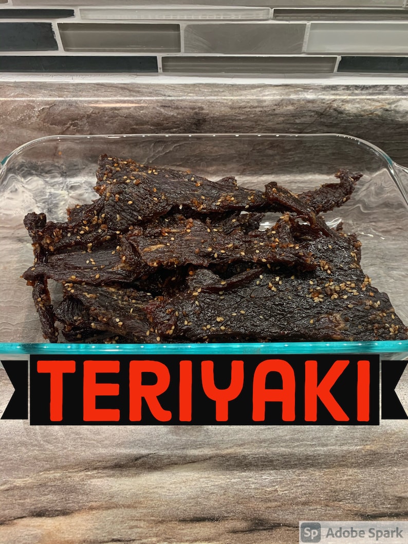 May include: A glass dish filled with dark brown, chewy, teriyaki beef jerky with sesame seeds. The dish is on a wooden surface with the word "TERIYAKI" in red letters on a black background.