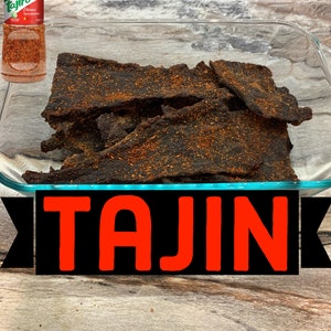 May include: A glass dish filled with dark brown beef jerky seasoned with Tajin Classic Seasoning. A bottle of Tajin Classic Seasoning is visible in the background.