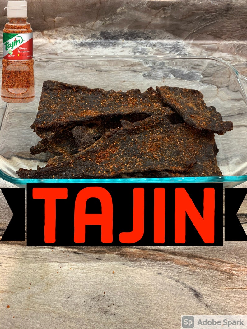 May include: A glass dish filled with dark brown beef jerky seasoned with Tajin Classic Seasoning. A bottle of Tajin Classic Seasoning is visible in the background.