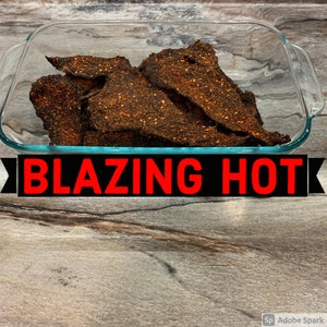 May include: A glass baking dish filled with a large amount of dark brown, spicy beef jerky. The dish is on a wood-grain surface. The text "BLAZING HOT" is printed in red letters on a black background.