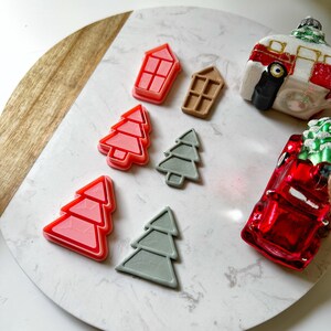 May include: A collection of Christmas-themed cookie cutters and ornaments. The cutters include red and gray Christmas trees and house shapes. Ornaments include a red truck and a camper. The items are arranged on a white and marble-patterned surface.