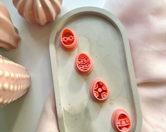 May include: Four pink Easter egg-shaped cookie cutters with different decorative designs are arranged on a light gray oval tray. The tray sits on a pink fabric surface, with pink ceramic decorative objects in the background.