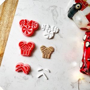 May include: A collection of Christmas-themed cookie cutters in various shapes. Includes red and white peppermint candy-shaped cutters, gingerbread house cutters, and other festive designs. The cutters are arranged on a white marble surface with holiday decorations.