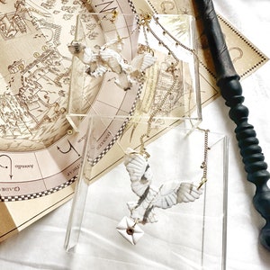 Puede incluir: Dos collares de búho blanco con cadenas doradas, cada uno con una pequeña carta. Los collares se exhiben sobre soportes de acrílico transparente, sobre un mapa y una varita negra. Los colgantes de búho tienen patrones de plumas detallados.