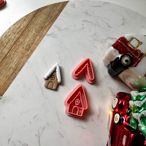 May include: A gingerbread house cookie and two pink plastic cookie cutters in the shape of a gingerbread house are displayed on a white and wood-grain surface. A red Christmas truck ornament is in the background.