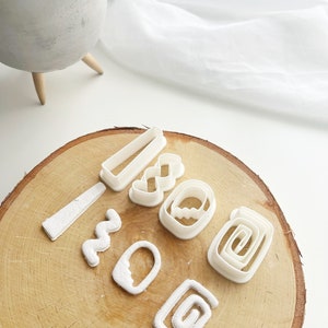 May include: A collection of white polymer clay cutters in various shapes, including rectangles, spirals, and wavy lines, arranged on a wooden surface. The cutters are for crafting and jewelry making.