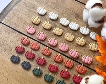May include: A collection of miniature pumpkin-shaped decorations in various colors, including white, tan, pink, orange, red, and green, with gold stems. The pumpkins are arranged on a wooden surface, with cotton and fall leaves.