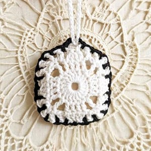 May include: A square crochet ornament featuring a white floral-inspired center pattern surrounded by a solid black border. The piece includes a white crocheted loop at the top for hanging, showcasing intricate yarn work and a contrasting two-tone design.
