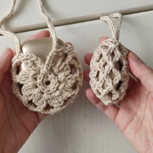 May include: Two beige crochet pouches made of thick cotton yarn. The larger pouch features a circular floral pattern and a long strap, while the smaller pouch has a diamond-shaped net design and a short loop. Both contain smooth gold objects.