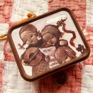 May include: A vintage, rectangular ornament with a brown wooden frame. The image inside depicts two children singing from sheet music, one playing a lute. The ornament has a gold cord for hanging and a black button on the back.