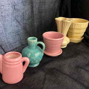 May include: A collection of ceramic vases and planters in various shapes and colors. Includes a pink teapot-shaped vase, a teal vase with a handle, a pink planter with a saucer, a cream-colored vase, and a stack of yellow planters.