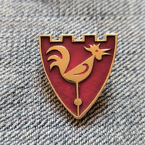 May include: A shield-shaped pin with a gold rooster design on a burgundy background. The pin has a gold border and a castle-like top. The rooster is facing right, with a detailed comb and tail. The pin is set against a denim background.