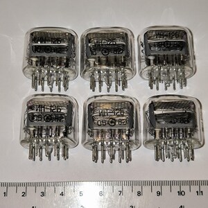 Peut inclure: Six tubes nixie en verre transparent disposés en deux rangées de trois, chacun avec des grilles métalliques internes et le texte cyrillique IN-12B. Une règle en bas montre que les tubes mesurent environ 3,2 cm de large, avec des broches.