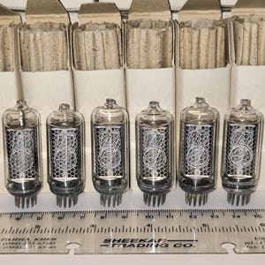May include: Six vintage vacuum tubes with clear glass bodies and intricate internal wire structures. Each tube is cylindrical with a base of pins. They are arranged in front of cardboard packaging.