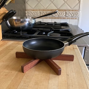 May include: A black frying pan with a wooden handle sits on a wooden trivet. The trivet has six arms that form a star shape.