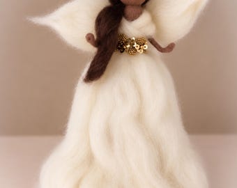 May include: A needle-felted angel figurine with dark brown wool hair, a cream-colored wool gown, and matching wings. The figure features a gold sequin headband and a gold sequin belt at the waist. The arms are thin and brown, extending outward.