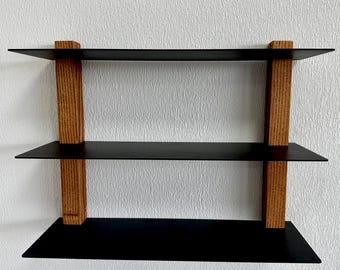 May include: A three-tiered wall shelf with a minimalist design. The shelves are black, and the supports are made of light brown wood. The shelf is mounted on a white wall, creating a contrast between the dark shelves and the light background.