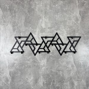 Op de afbeelding: Een zwarte metalen wandkapstok met een geometrisch ontwerp. De kapstok heeft een reeks onderling verbonden driehoekige vormen, wat een moderne en minimalistische uitstraling creëert. De kapstok is bevestigd aan een grijze muur met textuur.