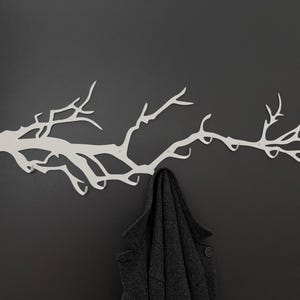 May include: A white metal wall-mounted coat rack shaped like a tree branch, mounted on a dark gray wall. A dark gray coat hangs from the rack, showcasing its functionality and design. The rack's design adds a touch of nature to the space.