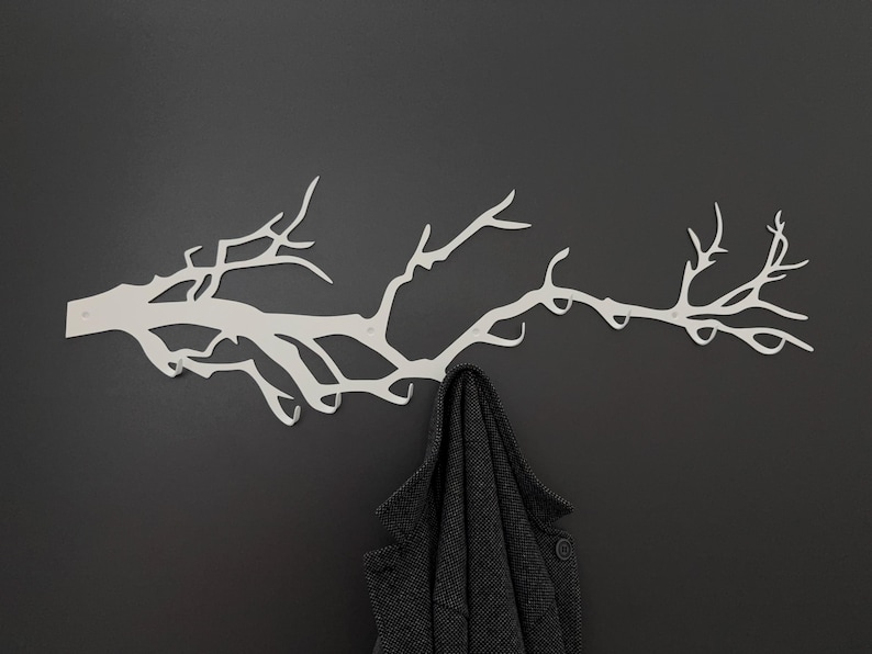 May include: A white metal wall-mounted coat rack shaped like a tree branch, mounted on a dark gray wall. A dark gray coat hangs from the rack, showcasing its functionality and design. The rack's design adds a touch of nature to the space.