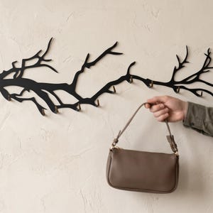 May include: A black metal wall-mounted coat rack shaped like a tree branch with multiple hooks. A person holds a brown purse with a shoulder strap. The rack is mounted on a textured, off-white wall.