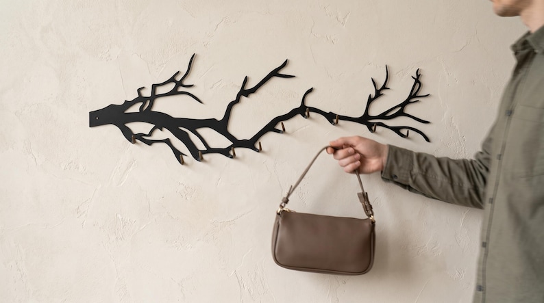 May include: A black metal wall-mounted coat rack shaped like a tree branch with multiple hooks. A person holds a brown purse with a shoulder strap. The rack is mounted on a textured, off-white wall.