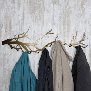 May include: A gold-colored metal wall-mounted coat rack in the shape of tree branches. Four coats in teal, gray, beige, and charcoal hang from the hooks. The rack is mounted on a white and gray wood-paneled wall.